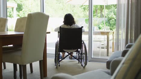 Biracial-disabled-woman-in-wheelchair-drinking-coffee-and-looking-out-of-window-in-sunny-living-room