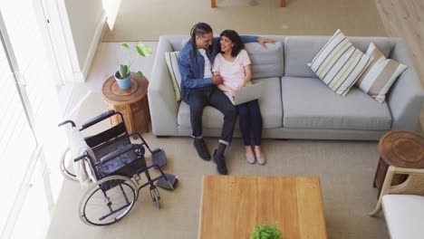Happy-biracial-couple-in-living-room-using-laptop-and-talking,-with-her-wheelchair-beside-couch
