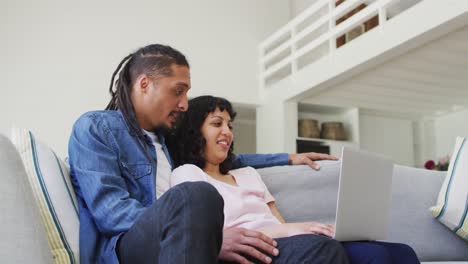 Happy-biracial-couple-sitting-on-couch-using-laptop-in-living-room