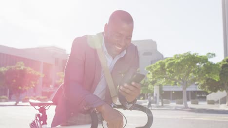 Hombre-Afroamericano-En-La-Ciudad,-Sentado-En-Bicicleta-Bajo-El-Sol-Usando-Un-Teléfono-Inteligente