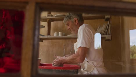 Senior-caucasian-man-wearing-apron-making-pottery-on-potter's-wheel-in-his-workshop