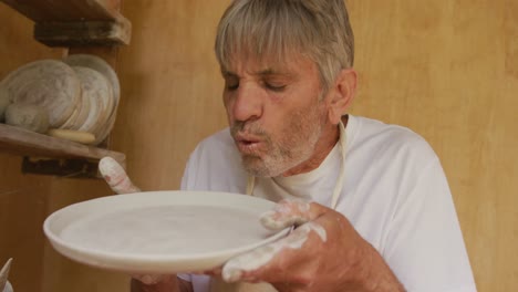 Senior-caucasian-man-wearing-apron-inspecting-clay-dish-in-pottery-workshop