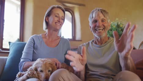 Portrait-of-smiling-senior-caucasian-couple-having-video-call-and-sitting-with-dog-in-living-room
