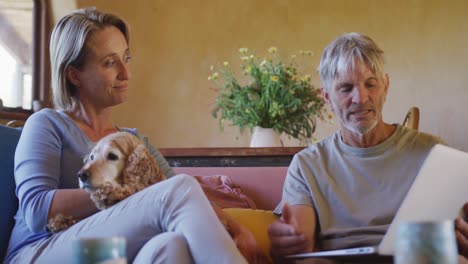 Happy-senior-caucasian-couple-sitting-in-living-room-at-home-with-pet-dog-using-tablet-and-talking