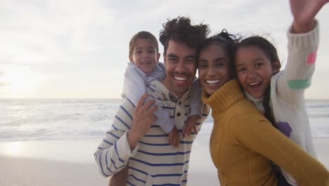 Portrait-of-happy-hispanic-parents-carrying-piggyback-children-on-beach