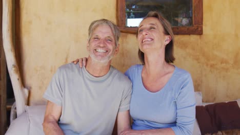 Portrait-of-smiling-senior-caucasian-couple-embracing-and-sitting-on-porch-outside-rustic-house