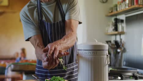 Senior-caucasian-man-wearing-apron-and-composting-vegetable-cuttings-in-kitchen