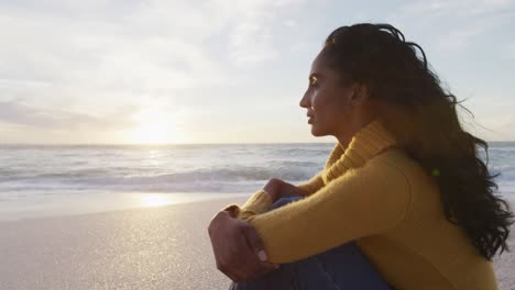 Portrait-of-thoughtful-hispanic-woman-sitting-on-beach-at-sunset