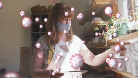 Animation-of-virus-cells-over-caucasian-woman-with-face-mask-inspecting-plants