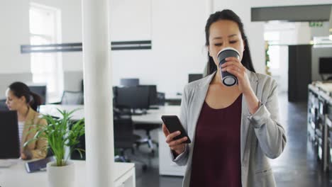 Happy-asian-businesswoman-walking-in-office,-holding-takeaway-coffee,-using-smartphone