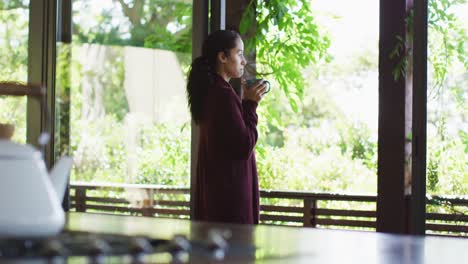 Relaxed-biracial-woman-drinking-coffee,-standing-at-window,-looking-into-distance
