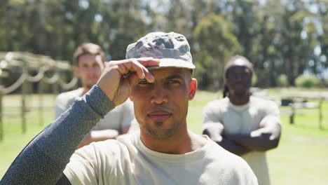 Portrait-of-african-american-male-soldier-in-cap-smiling-at-obstacle-course-with-two-men-behind-him