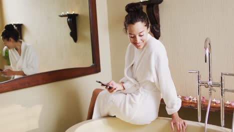 Glückliche-Biraziale-Frau-Mit-Vitiligo-Sitzt-Im-Bademantel,-Lässt-Die-Badewanne-Laufen-Und-Benutzt-Ihr-Smartphone