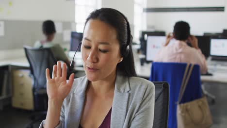 Happy-asian-businesswoman-sitting-at-desk-using-computer-and-talking-on-headset