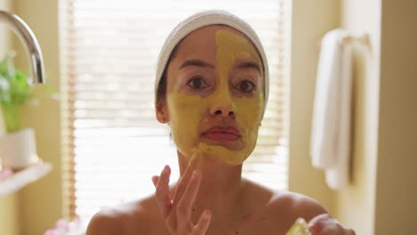 Portrait-of-biracial-woman-looking-into-mirror-and-applying-face-mask