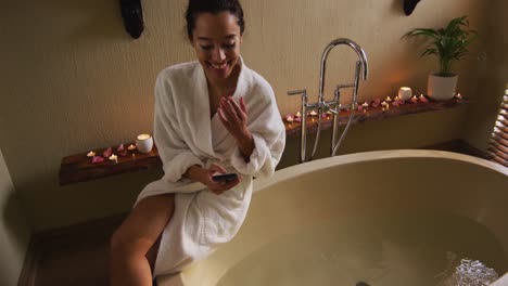 Happy-biracial-woman-with-vitiligo-sitting-in-bathrobe,-running-bath-and-using-smartphone