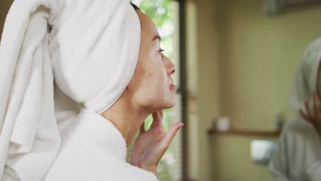 Biracial-woman-in-robe-looking-into-mirror-and-moisturizing-face