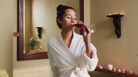 Relaxed-biracial-woman-with-vitiligo-wearing-bathrobe-and-drinking-wine-in-bathroom