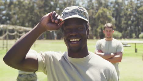 Portrait-of-smiling-african-american-male-soldier-in-cap-at-obstacle-course-with-others-behind-him