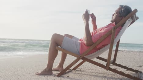 Latin-senior-hispanic-man-relaxing-on-sunbed-on-beach-at-sunset,-using-smartphone