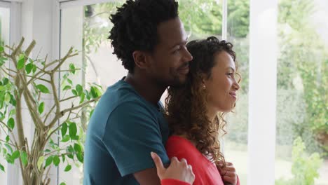Profiles-of-happy-biracial-couple-embracing,-standing-at-window-and-looking-into-distance