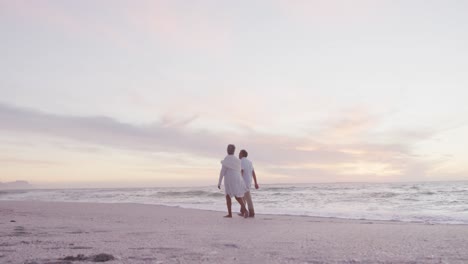 Back-view-of-hispanic-senior-couple-walking-on-beach-at-sunset