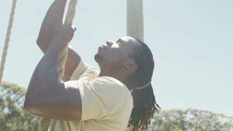 Fit-african-american-male-soldier-with-deadlocks-climbing-rope-on-obstacle-course-in-sun