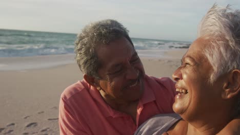 Portrait-of-happy-hispanic-senior-couple-laughing-and-embracing-on-beach-at-sunset