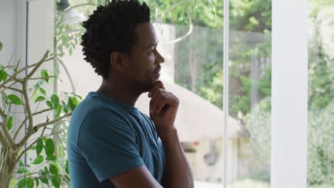 Thoughtful-biracial-man-standing-at-window-and-looking-into-distance