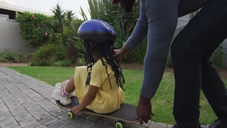 Happy-african-american-father-playing-with-daughter-on-skateboard-in-backyard