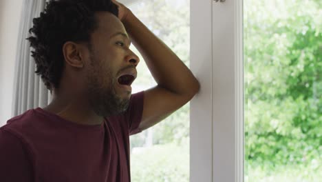 Profile-of-tired-biracial-man-standing-at-window-and-yawning