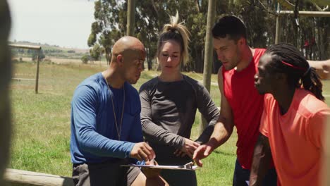 Diverse-fit-group-discussing-performance-with-male-trainer-at-obstacle-course-in-the-sun