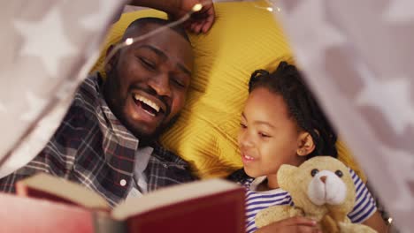 Happy-african-american-father-and-daughter-lying-in-tent-and-reading-book