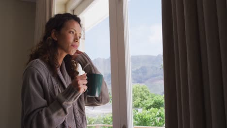 Thoughtful-biracial-woman-standing-with-coffee-at-window-and-looking-into-distance