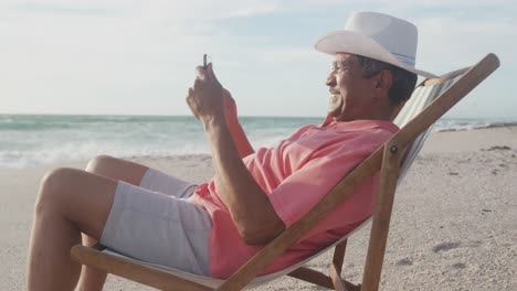 Feliz-Anciano-Hispano-Relajándose-En-Una-Tumbona-En-La-Playa-Al-Atardecer,-Usando-Un-Teléfono-Inteligente