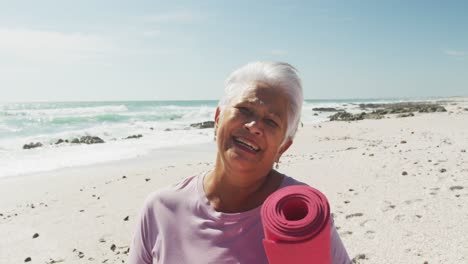 Portrait-of-hispanic-senior-woman-standing-on-beach,-holding-yoga-mat-and-smiling