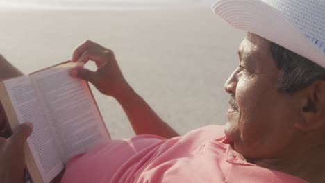 Profil-Eines-Hispanischen-älteren-Mannes,-Der-Sich-Bei-Sonnenuntergang-Auf-Einer-Sonnenliege-Am-Strand-Entspannt-Und-Ein-Buch-Liest
