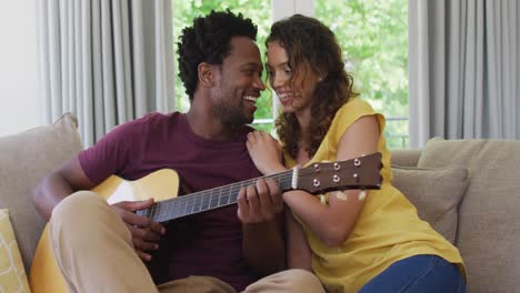Happy-biracial-couple-sitting-on-sofa-together-and-playing-guitar