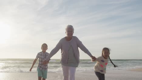 Glückliche-Hispanische-Großmutter-Und-Enkelkinder,-Die-Bei-Sonnenuntergang-Am-Strand-Laufen