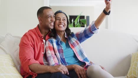Happy-biracial-couple-sitting-on-sofa,-embracing,-using-smartphone-and-laughing