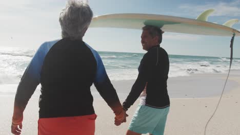 Back-view-of-happy-senior-hispanic-couple-walking-on-beach-with-surfboard