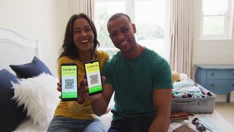 Happy-biracial-couple-sitting-on-bed-and-showing-smartphones-with-covid-19-passports-on-screens