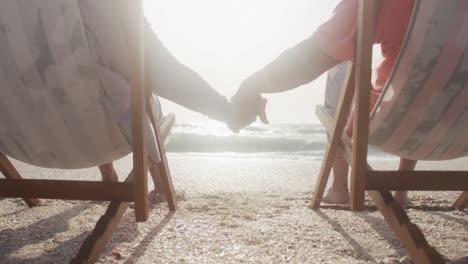 Back-view-of-hispanic-senior-couple-relaxing-on-sunbed-on-beach-at-sunset,-holding-hands