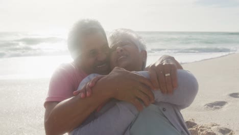 Happy-hispanic-senior-couple-sitting-and-embracing-on-beach-at-sunset