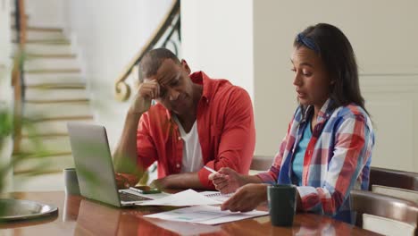 Stressed-biracial-couple-sitting-at-table-with-laptop-and-counting-home-budget