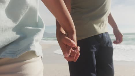 Midsection-of-hispanic-senior-couple-holding-hands,-walking-on-beach-at-sunset