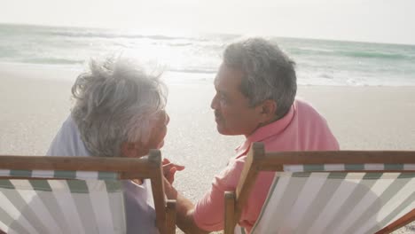 Happy-senior-hispanic-couple-kissing-and-relaxing-on-sunbeds-on-beach-at-sunset