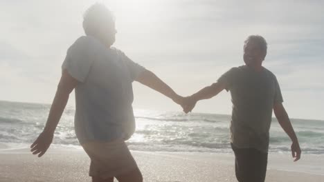 Happy-hispanic-senior-couple-holding-hands,-walking-on-beach-at-sunset