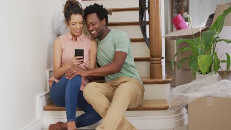 Happy-biracial-couple-taking-selfie-on-stairs-of-their-new-house