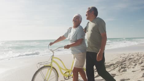 Happy-hispanic-senior-couple-walking-with-bikes-on-beach-at-sunset